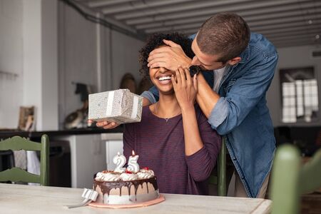 Young man giving present to his beloved girlfriend on birthday with cake on table. Cheerful african woman surprised by man during her 25th birthday. Guy covering girl eyes while surprising her with cake and present.の写真素材