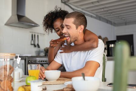 African american woman embracing from behind her boyfriend and feeding him with brown bread and jam. Multiethnic couple enjoying breakfast at home. Beautiful laughing woman feeding young man in kitchen and having fun together in the morning.の写真素材