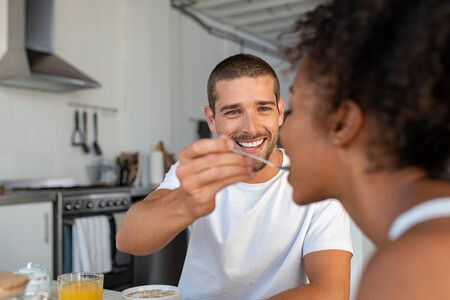 Happy multiethnic couple having breakfast in kitchen. Smiling handsome man feeding milk and cereal to woman in the morning. Girlfriend enjoying being pampered by boyfriend.の写真素材
