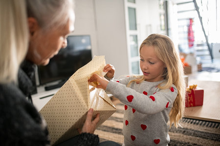Little girl wearing sweater and opening christmas present at home. Curious granddaughter open...