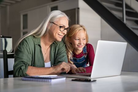 Happy grandchild and old grandmother sitting at home using laptop. Smiling cute girl and senior woman watching cartoons on laptop. Little granddaughter teach her beautiful granny how to use computer at homeの写真素材