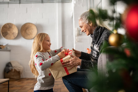 Happy senior woman giving christmas surprise to little girl at home. Excited granddaughter unpacking...