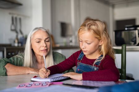 Beautiful girl child with senior woman writing on notebook. Cute little schoolgirl writing in notebook. Granddaughter with grandmother studying at home while sitting on table and doing homework together.の写真素材