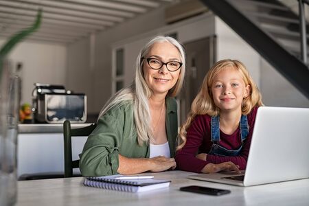 Portrait of cheerful granny with her beautiful smiling granddaughter looking at camera. Little girl teaching grandmother computer at home. Beautiful senior woman with cute grandchild sitting at table using notebook computer at home.の写真素材