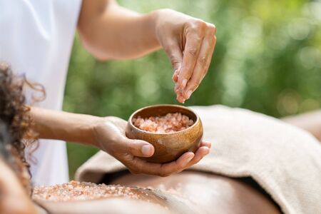 Closeup of woman lying and receiving massage with salt crystals at spa. Woman hand applying scrub on african woman back at wellness center for body exfoliation. Beauty therapist pouring salt for peeling on girl back.の写真素材