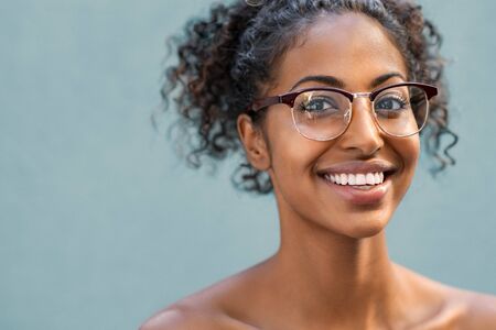 Cheerful young woman with naked shoulder wearing eyeglasses and looking at camera. Smiling african american woman with curly hair wearing spectacles isolated against blue background. Portrait of happy black girl with copy space.の写真素材