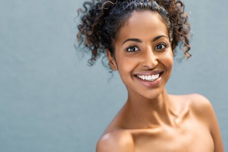 Smiling african american woman with curly hair looking at camera isolated over blue background with copy space. Happy black girl feeling good after skin treatment at spa. Wellness and wellbeing concept.の写真素材