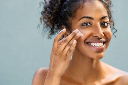 Beautiful african american woman with smooth skin applying moisturizer face cream to her cheek. Beauty young woman taking care of skin. Happy girl applying cosmetic moisturiser treatment isolated on background and looking at camera with copy space.の写真素材