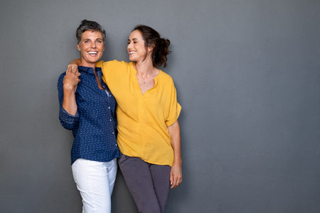 Mature happy women embracing each other against grey wall with copy space. Happy laughing ladies in smart casual standing on gray background. Cheerful middle aged woman with hand on shoulder of her stylish friend.の写真素材
