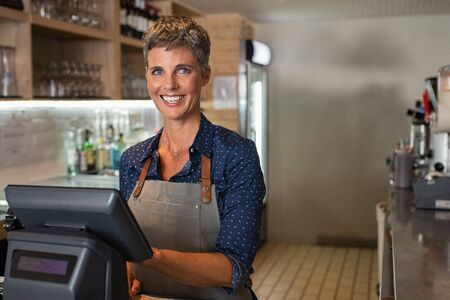 Happy waitress wearing apron standing by a point of sale terminal and laughing while working in a coffee shop. Mature woman entrepreneur standing at  counter operating computer to prepare bill for customer. Portrait of satisfied and successful small business owner standing while looking at camera.の写真素材