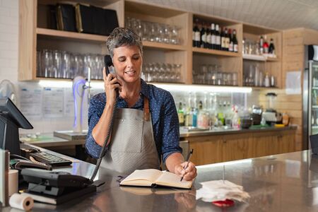Barista woman taking orders over the phone at counter. Mature woman in grey apron writing on customer book while talking over phone in cafeteria. Waitress writing takeaway lunch while using telephone.の写真素材