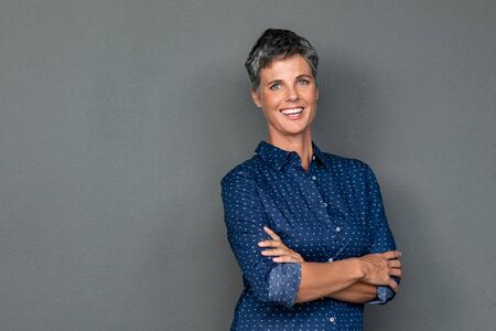Isolated portrait of successful businesswoman with crossed arms looking at camera. Portrait of happy mature woman standing with arms folded isolated over gray background. Confident business woman smiling with copy space.の写真素材