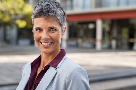 Portrait of smiling mature businesswoman in formal clothing standing outdoor and looking at camera. Successful senior entrepreneur looking at camera on street. Confident corporate manager smiling and feeling proud with copy space.の写真素材