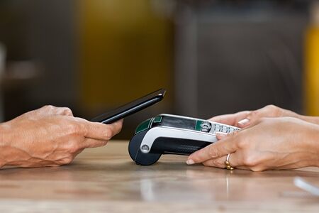 Closeup of woman hand paying a bill through smartphone using NFC technology. Closeup of hand holding pos terminal to receive payment using mobile phone. Customer using mobile to make payment with contactless.の写真素材