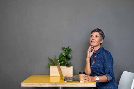 Thoughtful mature businesswoman looking away while using laptop at table in creative office. Beautiful senior woman working on computer and looking up on gray wall while thinking about the future. Smiling pensive business lady isolated against grey background contemplating plans.の写真素材