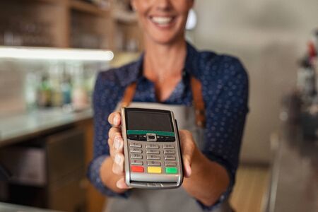 Waitress standing at cash counter holding an electronic card payment machine. Close up of mature woman hand holding wireless terminal POS at coffee shop. Happy smiling owner showing NFC technology machine to customer across counter to make payment.の写真素材