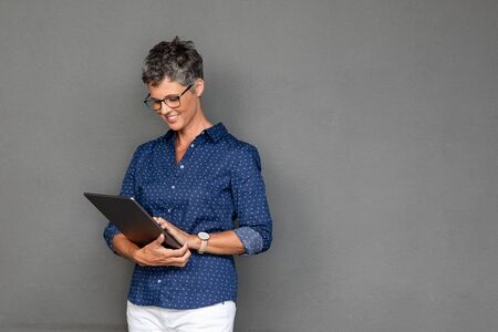 Smiling mature businesswoman wearing spectacles while using digital tablet against grey background with copy space. Successful senior business woman surfing internet on digital tablet on gray wall. Beautiful middle aged woman checking email on laptop.の写真素材