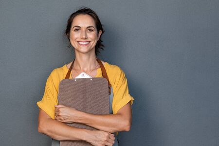 Portrait of smiling mature woman in apron holding clipboard in hand while looking at camera. Beautiful waitress standing isolated against grey wall with copy space. Successful small business owner standing on grey background with copy space.の写真素材