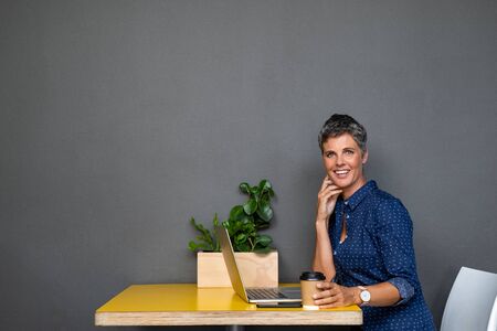 Successful business woman sitting at coffee shop working on laptop while drinking hot coffee. Mature woman holding take away coffee cup while using computer and looking at camera. Woman entrepreneur sitting at table with grey background and copy space.の写真素材