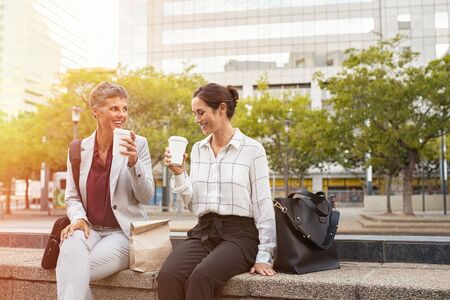 Smiling business partners having break outdoor sitting in a square. Happy mature woman and her colleague enjoying and drinking coffee in city centre. Two smiling women relaxing in conversation outside office with copy space.の写真素材