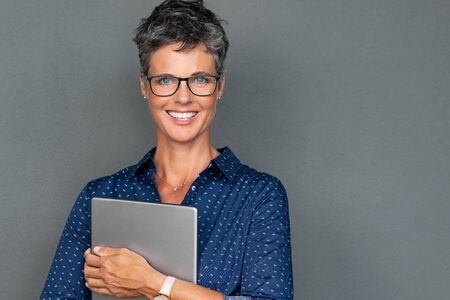 Mature stylish woman wearing eyeglasses and smiling while holding digital tablet. Beautiful senior woman looking to camera isolated on grey background. Portrait of businesswoman with grey hair and spectacles standing  with copy space.の写真素材
