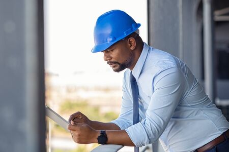 Mature manager using digital tablet on construction site. Serious african engineer checking email at building site wearing blue hardhat. Architect working on digital tablet leaning at balustrade.の写真素材