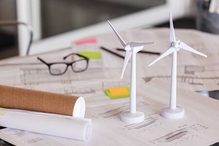 Close up of wind turbines on a table with architectural plan blueprints. Two models of wind turbines on desk with architecture layout and paper. Green and eco sustainable architecture.の写真素材