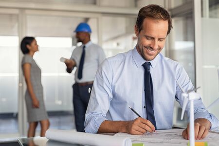 Architect man working on blueprints for new eco sustainable construction. Smiling mature architect at work in the studio with copy space. Engineer working on wind turbines implantation with team in background.の写真素材