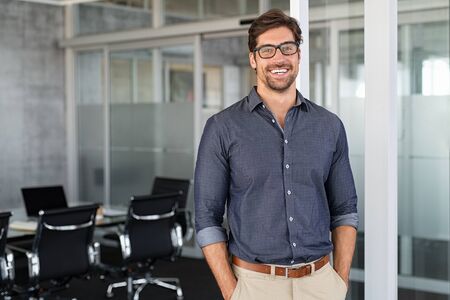 Portrait of young businessman wearing eyeglasses and standing outside conference room. Portrait of happy business man wearing spectacles and looking at camera with copy space. Satisfied proud man feeling confident in a modern office.の写真素材
