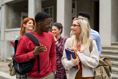 Group of happy multiethnic students walking outside college building. Five university students holding books and school backpack while talking together. Young man and beautiful girl in conversation at college campus.の写真素材
