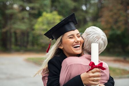 Enthusiastic graduated daughter holding degree embracing mother in campus. Young female student graduate hugging her mother at graduation ceremony. Excited college student with the graduation gown and hat holding diploma and hug the parent at campus.の写真素材