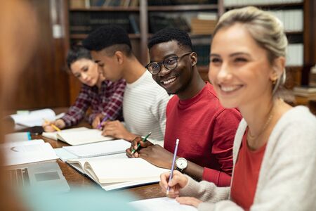 Group of happy multiethnic students studying and writing together in a library. College students in a library smiling. Young men and women sitting in a row at university library.の写真素材