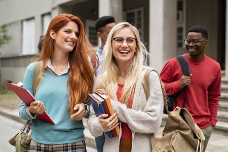 Multiethnic group of happy young women and men walking in college campus. Cheerful group of multiethnic friends holding books and feeling relaxed after university exam. Smiling classmates laughing and looking away.の写真素材