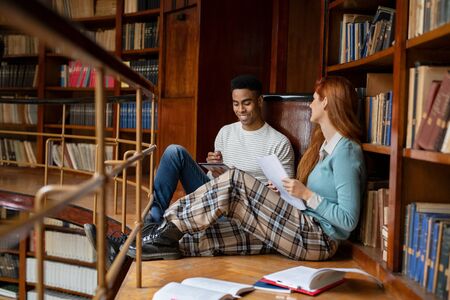 Couple of university students at library preparing exams sitting on library floor. Happy friends studying together at college library. Smiling young woman and african man getting ready for exams together. の写真素材