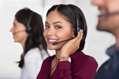 Smiling beautiful woman working in call center as telemarketing operator. Smiling customer support agent wearing headset at office, working while looking at camera. Middle eastern girl working as telephone operator with her colleagues.の写真素材