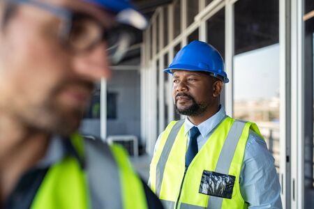 Engineer builder at construction site looking away and thinking. Mature african man architect wearing safety helmet, contemplating construction site. Thoughtful construction worker daydreaming, vision concept.の写真素材