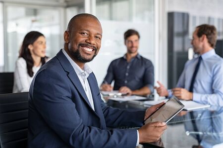 Mature black businessman with colleagues sitting in a modern board room. Proud smiling business man sitting during a meeting and looking at camera. Portrait of happy successful executive with team working in background.の写真素材