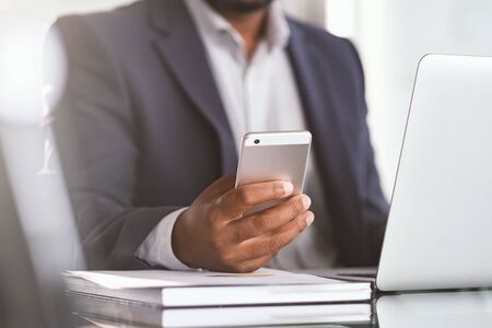 Close up of black businessman hand holding smartphone while working on laptop in modern office. Closeup of african hand using cellphone at desk. Mature man typing on mobile phone while working.の写真素材