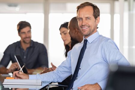 Portrait of smiling businessman at conference with his team in background. Successful business man looking at camera in the meeting room while colleagues working behind. Handsome middle aged formal man with business people in background at modern office.の写真素材