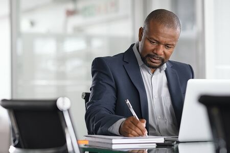 African serious businessman writing notes and using laptop. Mature business man writing his strategy on notebook while using laptop in modern office. Focused black entrepreneur sitting at desk in modern office while working.の写真素材