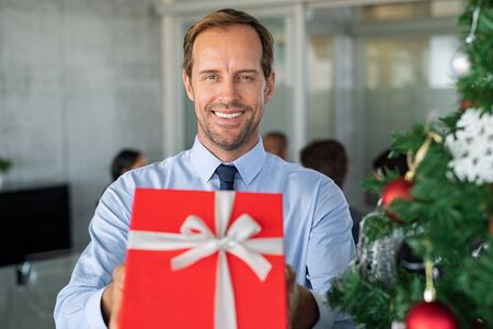 Successful mature business man holding a red gift box near christmas tree in modern office. Portrait of cheerful businessman presenting surprise gift in meeting. Man in formal clothing offer a xmas gift while looking at camera with a big grin.の写真素材