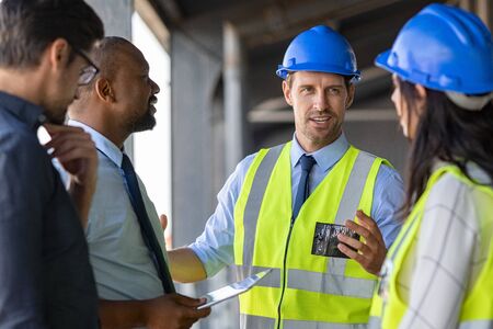 Happy mature engineer discussing the structure of the building with architects colleague at construction site. Engineers wearing safety hardhat having work conversation on the safety of the structure. Multiethnic team of business men and workers talking at site.の写真素材