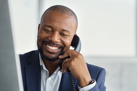 Close up face of mature businessman working on computer and talking on phone. Mature african man talking on mobile phone at desk in office. Smiling manager working at desk with copy space.の写真素材