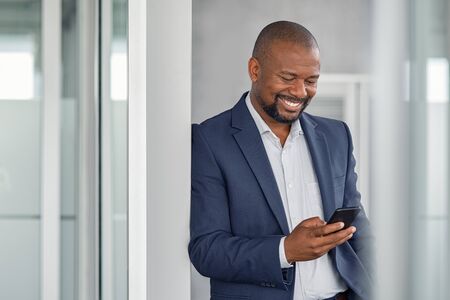 Happy mature businessman in formal using smartphone in modern office. Successful african american business man smiling while reading message on cellphone. Middle aged man in formal using mobile phone in modern workplace.の写真素材