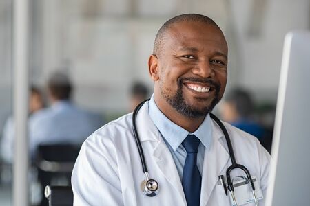 Happy smiling african doctor looking at camera in medical office. Portrait of black man doctor working on laptop in modern hospital. Confident and reliable mature nurse using computer to analyze medical report of patient.の写真素材