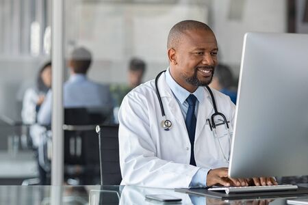 Practitioner typing on computer in modern clinic. Doctor at work in office using computer with copy space. African happy specialist wearing white coat and sitting at desk while mediacal team having meeting in background.の写真素材