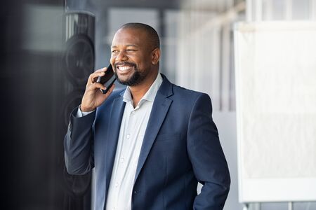 Successful black businessman in a happy conversation over cellphone. Mature african american business man in formal suit talking over phone in moder office. Happy man calling on smartphone while looking through the window.の写真素材