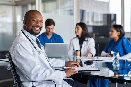 Portrait of cheerful doctor looking at camera and smiling with working clinicians behind. Mature african doctor and medical staff working in hospital. Successful clinic worker in labcoat and stethoscope sitting in conference room with copy space.の写真素材