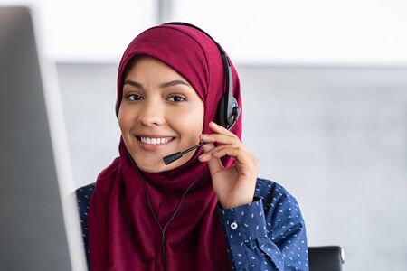 Portrait of happy young islamic woman in headscarf holding microphone and working on computer. Arab businesswoman working in customer service center talking using headset. Portrait of beautiful arabic woman in hijab smiling and looking at camera while working in office with copy space.の写真素材