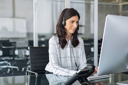 Mature secretary working in call center with headset. Confident telephone operator in modern office working on desktop computer wearing headset while taking calls. Smiling customer support operator at work with copy space.の写真素材